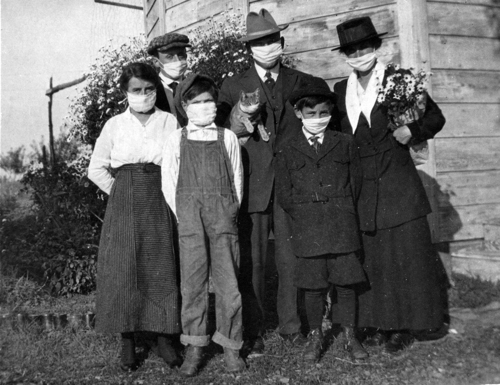 A caucasian family of three adults, three kids, and a cat pose outdoors of their home wearing white masks in a black and white photo